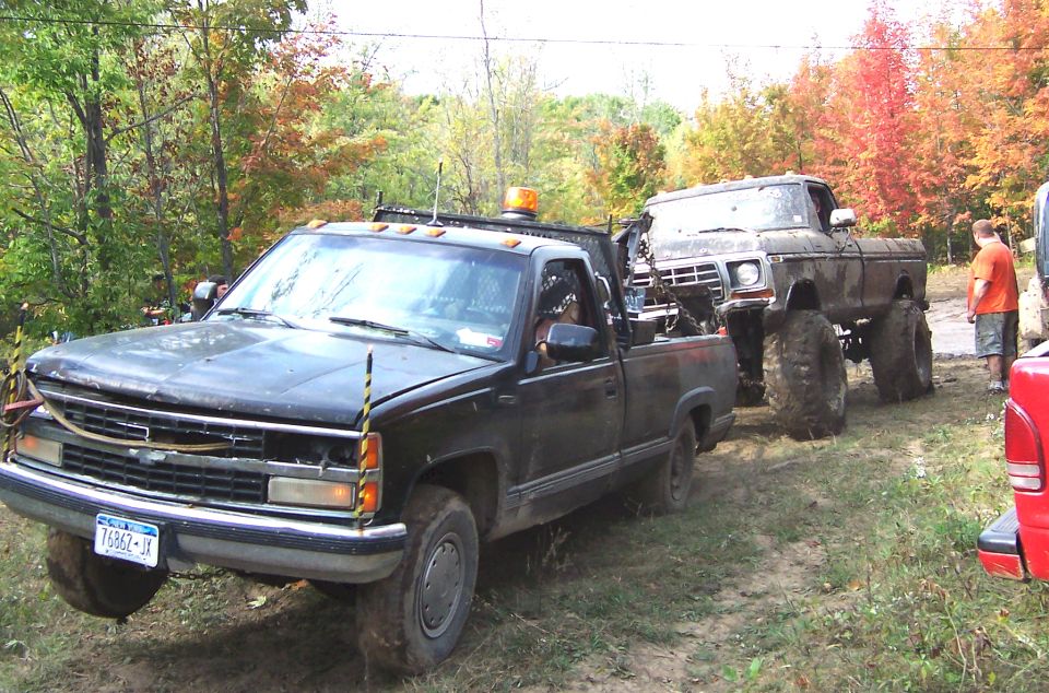 Muddy lifted Ford truck being towed by a black Chevrolet pickup; red vehicle visible on right