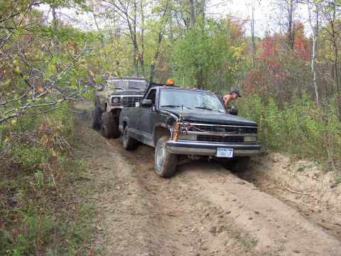 1978 Ford Bronco - Two off-road vehicles navigate a challenging dirt path through a wooded area.