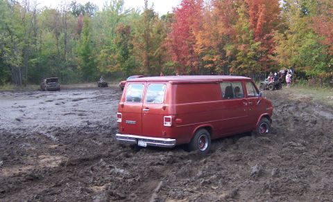 1978 Ford Bronco - A red van is stuck in a muddy area with trees and other vehicles in the background.