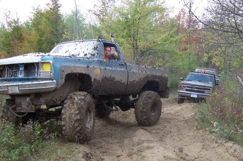 1978 Ford Bronco - Two heavily modified trucks navigate a muddy off-road path through a wooded area.