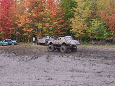 1978 Ford Bronco - A monster truck and other vehicles are in a muddy field with fall foliage in the background.