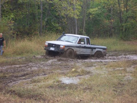 1978 Ford Bronco - A pickup truck navigates a muddy off-road trail, with trees and grass in the background.
