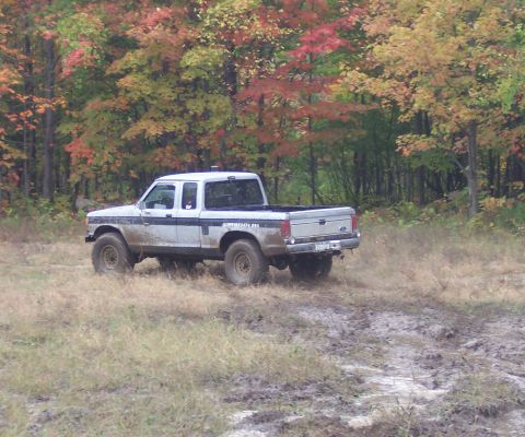 1978 Ford Bronco - Side view of a two-tone white and blue Ford F-Series extended-cab 4x4 pickup parked on grass beside a mud pit, its lower body and steel whe…