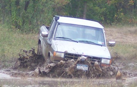 1978 Ford Bronco - Close front three-quarter view of a white Ford pickup with a body-mounted snorkel churning through a soupy mud hole, clods of mud flying up…