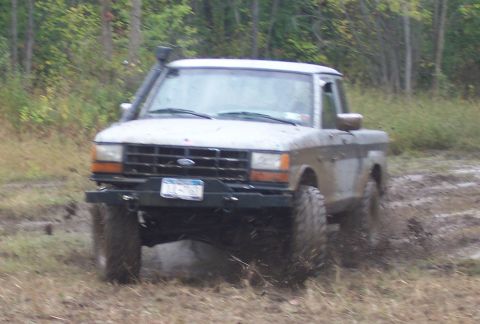 1978 Ford Bronco - A mud-caked white Ford F-Series regular-cab pickup, lifted on aggressive mud-terrain tires, drives out of a churned mud hole at the edge of…