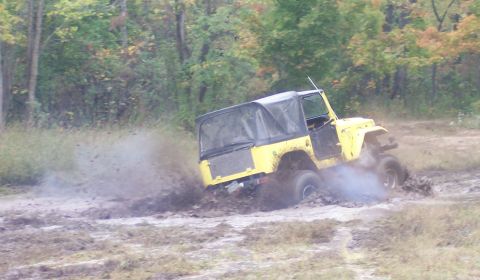 1978 Ford Bronco - Rear three-quarter view of a yellow Toyota Land Cruiser FJ40 with a black soft top digging through a mud pit, rooster tails of muck flying…