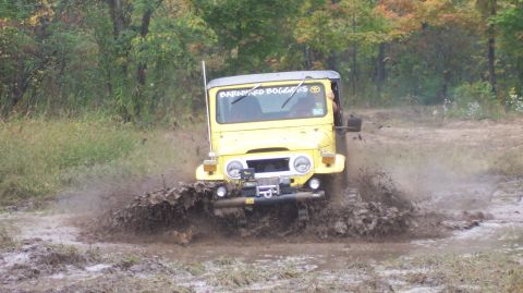 1978 Ford Bronco - Head-on view of a yellow Toyota Land Cruiser FJ40 blasting through a wide mud pit, water and muck spraying off both front tires. The truck…