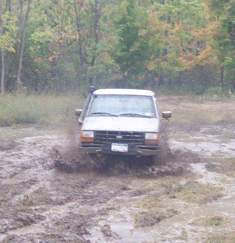 1978 Ford Bronco - Head-on shot of a white Ford truck with a snorkel running up the passenger A-pillar plowing through a deep, water-filled mud pit, brown spr…