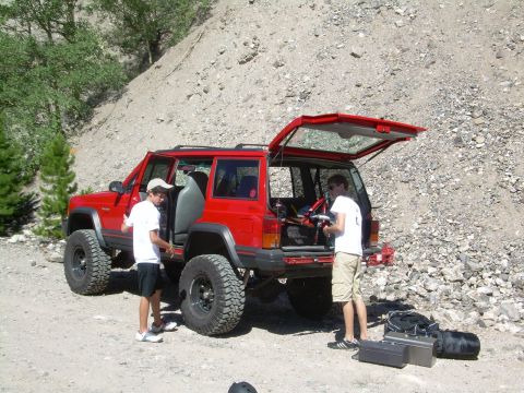 1996 Jeep Cherokee - Two people load gear into a red Jeep Cherokee parked on a dirt road with a rocky hillside in the background.