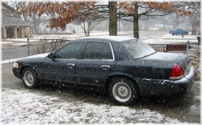 1999 Ford Crown Victoria - A dark sedan sits parked on a snow-covered street during a snowfall.