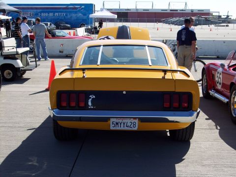 1971 Ford Mustang - Rear view of a yellow car with a black stripe on the hood and trunk, parked outdoors with other vehicles.
