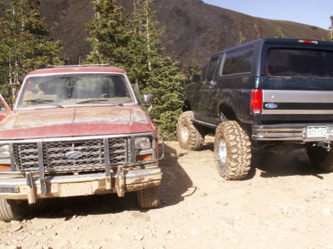 1986 Ford Bronco - Two modified Ford Broncos are parked on a dirt road in a mountainous, wooded area.