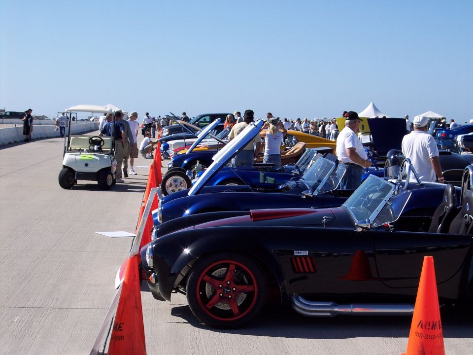 Several classic sports cars are parked in a line on a paved surface under a clear blue sky. A golf cart is visible to the left