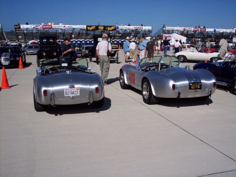 1971 Ford Mustang - Two classic Shelby Cobra sports cars are displayed at an outdoor event with spectators in the background.