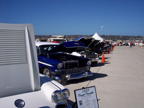 1971 Ford Mustang - Several classic cars are lined up with their hoods open at an outdoor event, with a sign for a 1958 Corvette Convertible in the foreground.