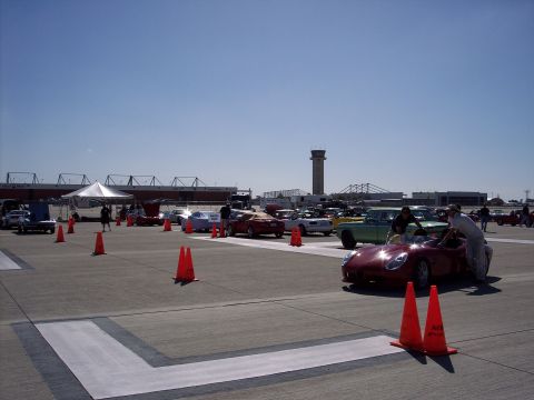 1971 Ford Mustang - Several cars are parked on an airfield under a clear blue sky, with a control tower in the distance.