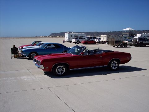 1971 Ford Mustang - A red convertible classic car is parked on a paved lot under a clear blue sky with other vehicles and trailers in the distance.