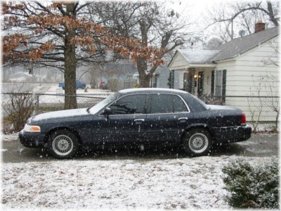 1999 Ford Crown Victoria - A dark sedan sits on a snow-covered road in front of a house during a snowfall.