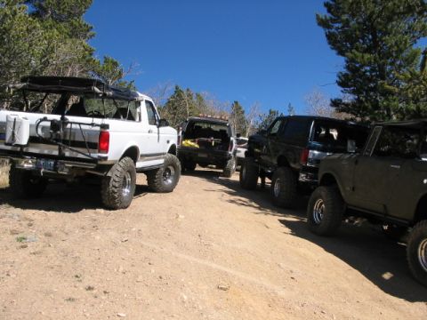 1995 Ford Bronco - A group of off-road vehicles, including a white 1995 Ford Bronco, are parked on a dirt road under a clear blue sky.
