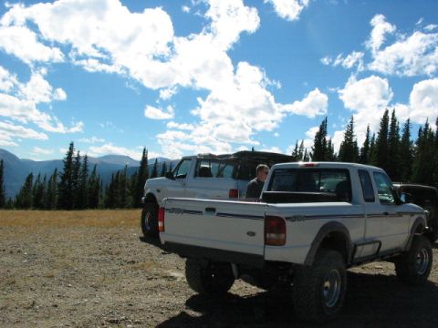 1995 Ford Bronco - A scenic mountain landscape with several vehicles parked on a dirt road under a cloudy sky.
