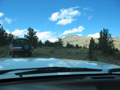 1995 Ford Bronco - Two vehicles on a dirt road in a mountainous landscape under a blue sky with clouds.