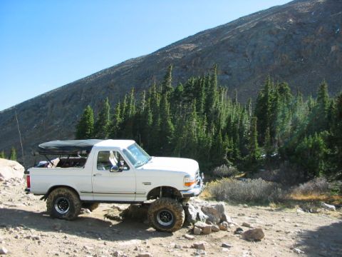1995 Ford Bronco - A lifted white Ford Bronco is parked on a rocky trail with a steep mountain and pine forest behind it.