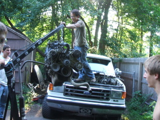 1989 Ford Bronco - A man uses a crane to lift an engine from the front of a white Ford truck, with others watching.