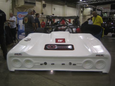 1990 Ford Bronco - white fiberglass off-road race body shell with headlight cutouts sits on display at an indoor show with onlookers behind.