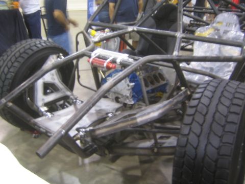 1990 Ford Bronco - bare tube-frame off-road race chassis with a mounted engine block and red coilover shock sits on display at an indoor show.
