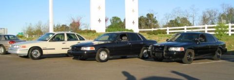 1999 Ford Crown Victoria - Three Ford Crown Victoria police cars are parked in a lot. The tan car is on the left, followed by two black cars.