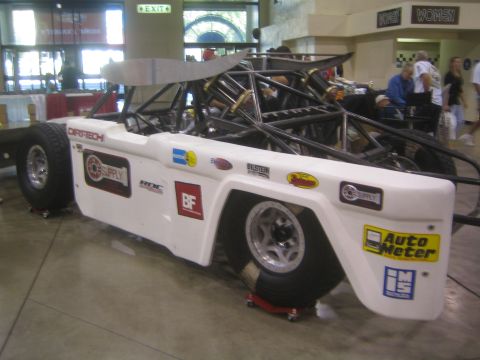 1990 Ford Bronco - rear quarter of a white tube-frame off-road race truck shows Dirt-Tech, Auto Meter and other sponsor decals at an indoor show.