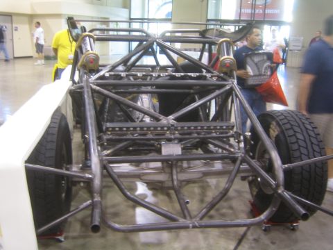 1990 Ford Bronco - rear of a bare welded tube-frame off-road chassis sits on dollies at an indoor show as a person holds a red body panel nearby.