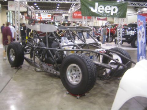 1990 Ford Bronco - tube-frame off-road race buggy sits on dollies beneath a green Jeep banner at an indoor automotive show.