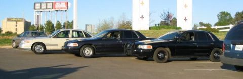 1999 Ford Crown Victoria - A lineup of police cruisers, including a tan model and two black models, are parked in a lot under a bright sky.