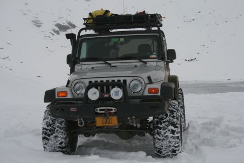 2003 Jeep TJ - A silver Jeep Wrangler is parked in a snowy landscape, equipped with a snorkel and a roof rack loaded with gear.