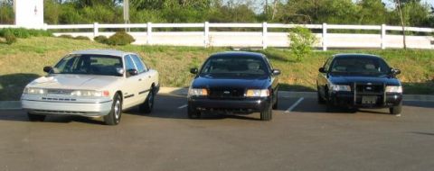 1999 Ford Crown Victoria - Three Ford Crown Victorias are parked in a lot, with a light-colored sedan in the foreground and two dark sedans behind it.