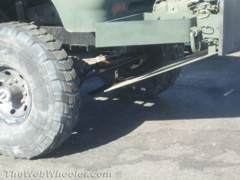 1990 Ford Bronco - Low-angle undercarriage shot of an olive-drab 4x4 truck showing a heavy off-road tire, axle, frame and side step rail.