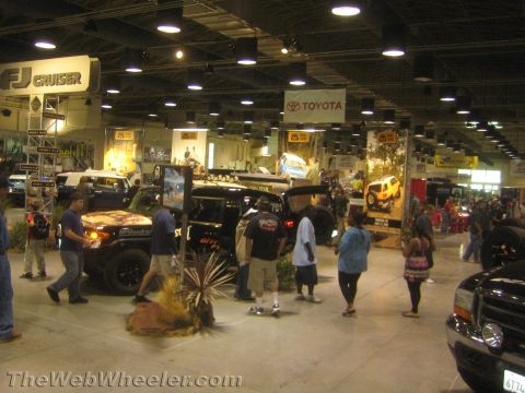 1990 Ford Bronco - Crowds walk an indoor automotive expo floor lined with lifted off-road trucks under FJ Cruiser and Toyota banners.