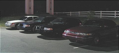 1999 Ford Crown Victoria - A line of sedans, including police-style vehicles, parked in a lot at night.
