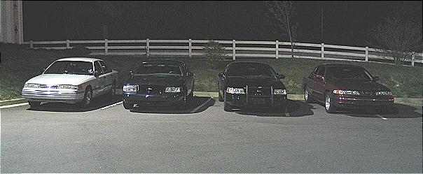 four sedans parked in a lot at night, one white and three dark colored