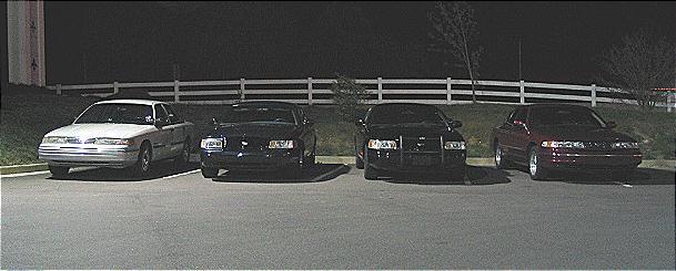 Four cars parked in a lot at night. The first car is white, the other three are dark. The 1999 Ford Crown Victoria is visible