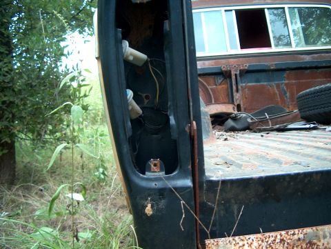 1979 Ford Bronco - A close-up view of a heavily rusted truck bed and its missing tail light assembly, surrounded by vegetation.