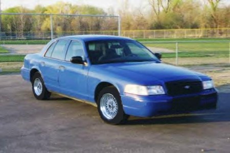 1999 Ford Crown Victoria - A blue sedan is parked outside on a paved surface with a fence and trees in the background.