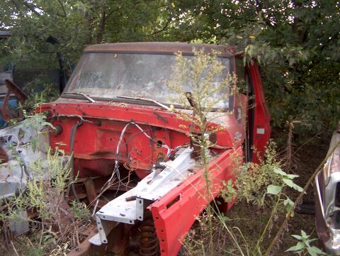 1979 Ford Bronco - Front view of a derelict red truck cab in a junkyard, surrounded by vegetation.
