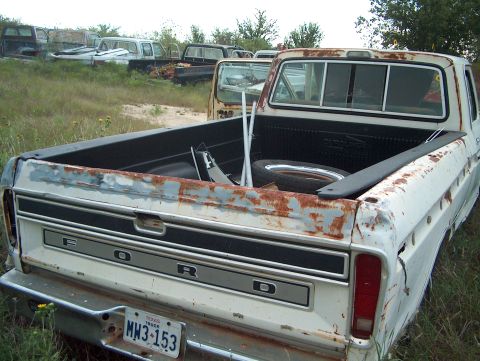 1979 Ford Bronco - A rusty white Ford pickup truck sits in a junkyard with other vehicles in the background.