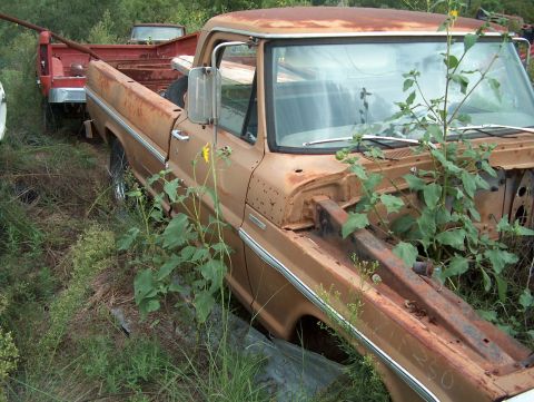 1979 Ford Bronco - This rusty brown pickup truck, likely from the late 1970s, sits in a junkyard with weeds growing through it.