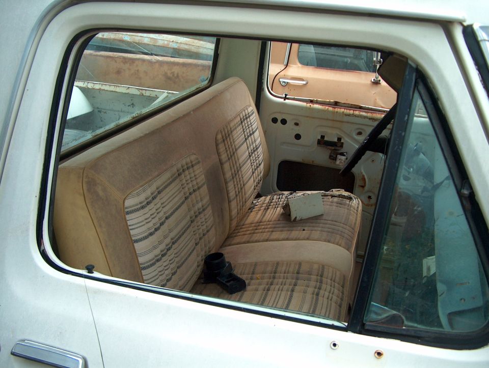 Interior view of a white pickup truck showing a plaid bench seat, door panel removed, and another vehicle visible through the window