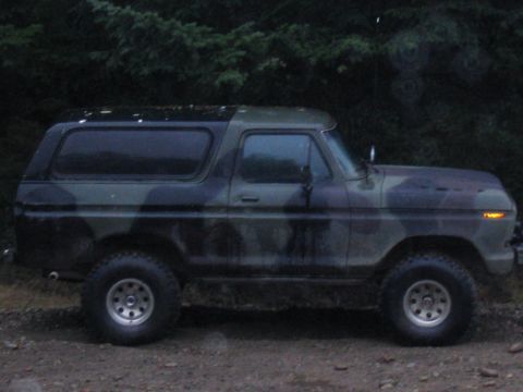 1989 Ford Bronco - A camouflaged Ford Bronco is parked on a dirt road with trees in the background.