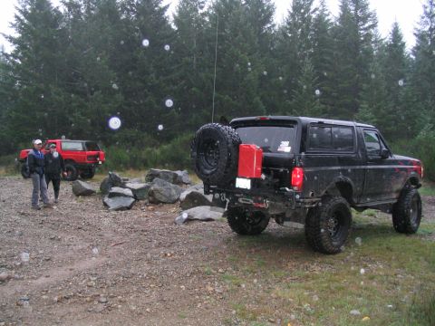 1989 Ford Bronco - Two off-road vehicles are parked in a wooded area with rocks scattered around.