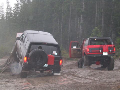 1989 Ford Bronco - Two modified Broncos navigate a wet, wooded trail with one vehicle partially on a log.